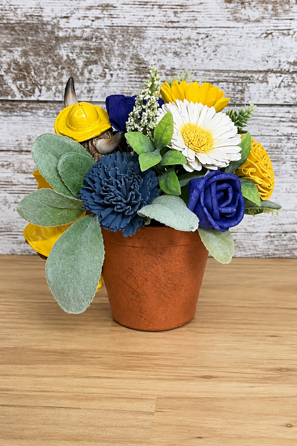Royal blue and slate blue sola wood blooms with white daisies, yellow flowers, and soft sage foliage in a brown planter