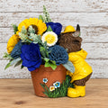 Blue, yellow, and white sola wood flowers beside a Highland cow in yellow rainwear on a brown floral planter