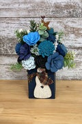 A decorative faux floral arrangement in a dark blue rectangular vase with a rustic wooden reindeer decoration and plaid bow, displayed on a weathered wood plank background.