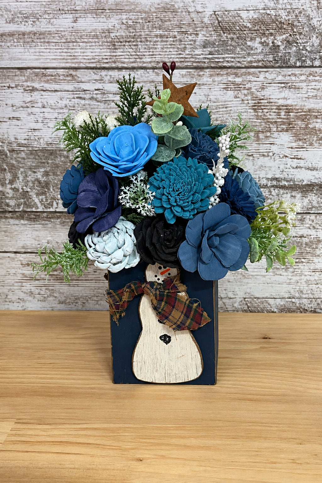 A decorative faux floral arrangement in a dark blue rectangular vase with a rustic wooden reindeer decoration and plaid bow, displayed on a weathered wood plank background.